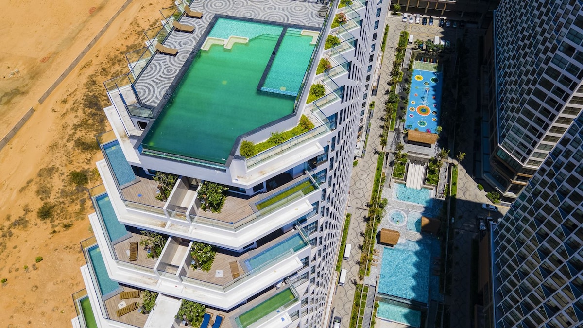 An aerial view captures a modern multi-level building featuring several swimming pools, each surrounded by lush greenery. The pools are arranged in a tiered design, creating a sense of spaciousness and inviting leisure in a vibrant environment.