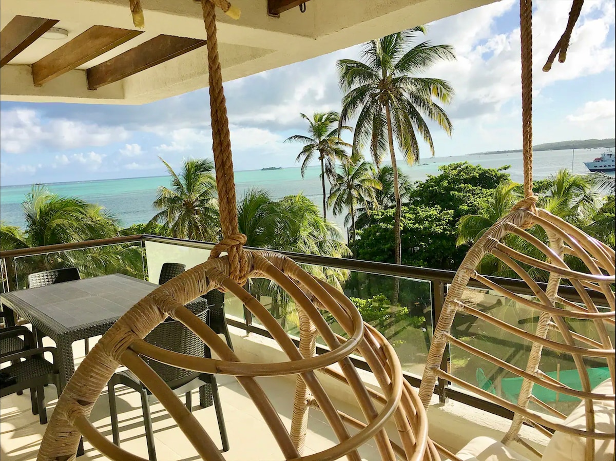 A spacious balcony features two hanging chairs and a dining table, surrounded by palm trees. The serene view of the ocean stretches toward the horizon, with gentle waves lapping against the shore, creating a relaxing environment.