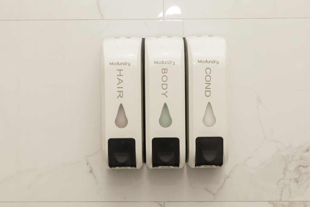 Three wall-mounted dispensers are neatly arranged, labeled for hair, body, and conditioner. The sleek white containers contrast against the light marble background, providing an organized and modern touch to the bathroom's design.