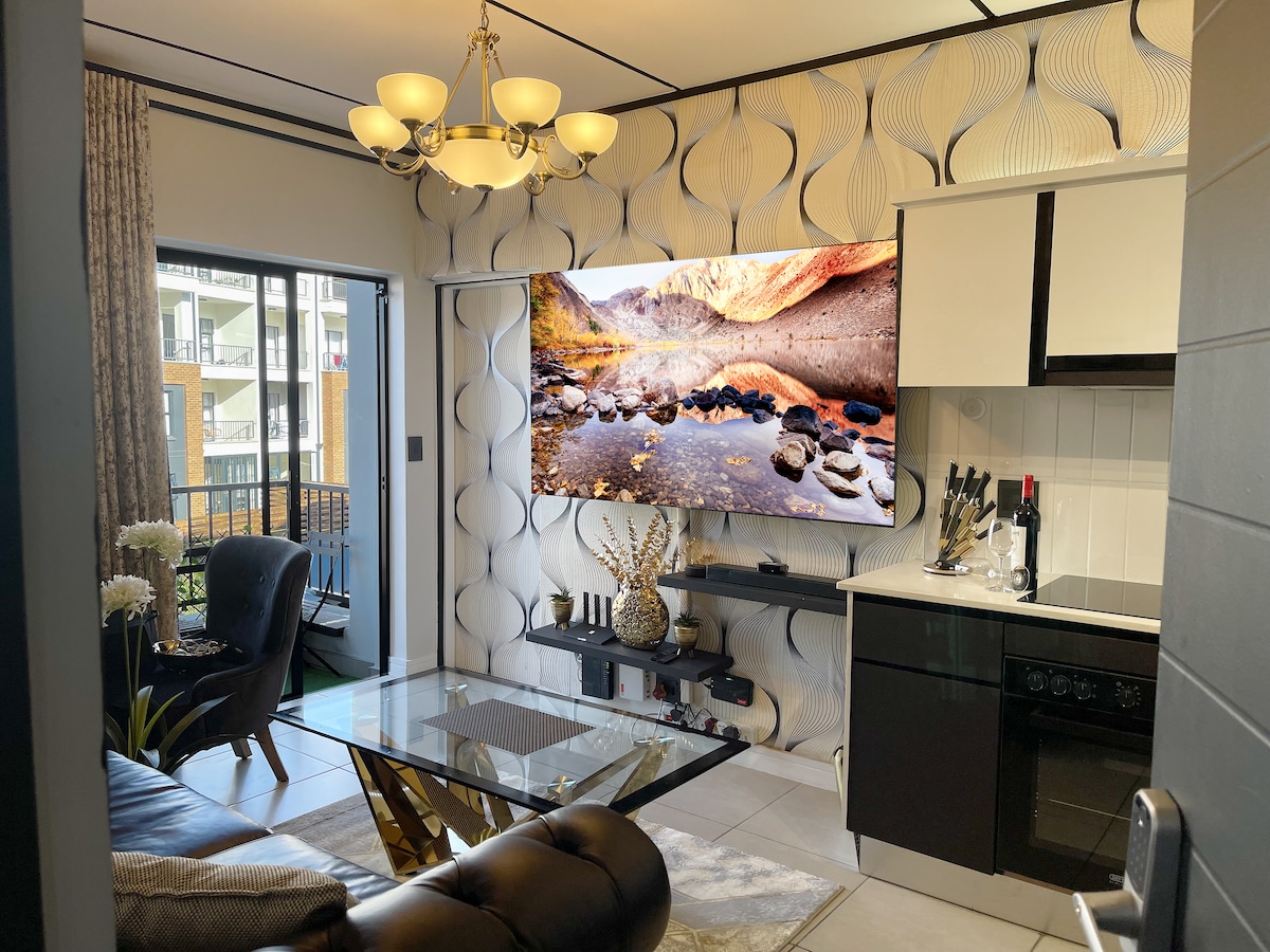 A contemporary living area is illuminated by a stylish chandelier, featuring a large wall-mounted TV displaying a scenic landscape. A sleek glass coffee table sits in front of a plush leather couch, complemented by decorative elements and a light-colored kitchen area visible in the background.