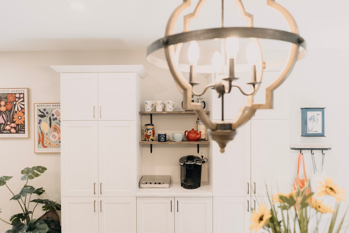 A modern kitchen is showcased with white cabinetry and a contrasting dark countertop. A coffee maker is positioned on the counter beneath open shelving displaying various decorative items. A chandelier hangs above, adding elegant illumination, while a potted plant and framed artwork provide warm accents.