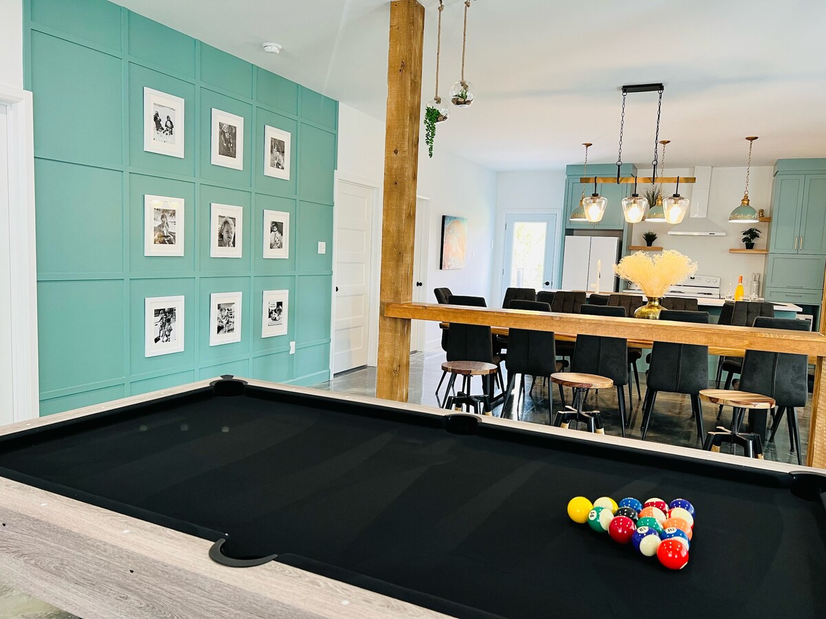 An interior view of a modern chalet showcases a pool table in the foreground, with a set of colorful balls arranged for a game. A dining area with eight black chairs and a wooden table is visible in the background, complemented by teal accent walls and framed artwork.