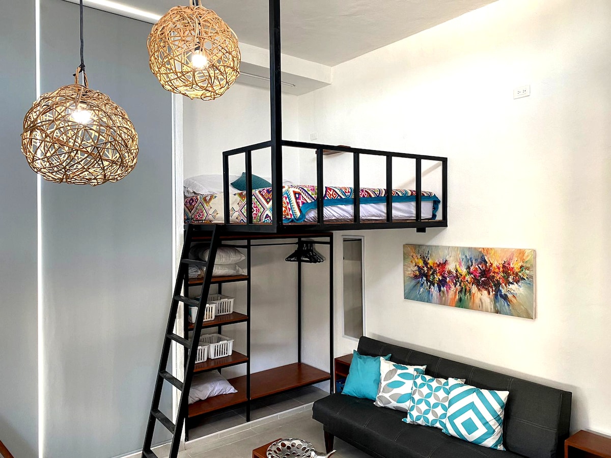A modern living space features a lofted sleeping area accessed by a ladder. Below, a dark sofa is complemented by colorful cushions. A light-hued abstract painting hangs on one wall, while decorative hanging lights provide illumination.