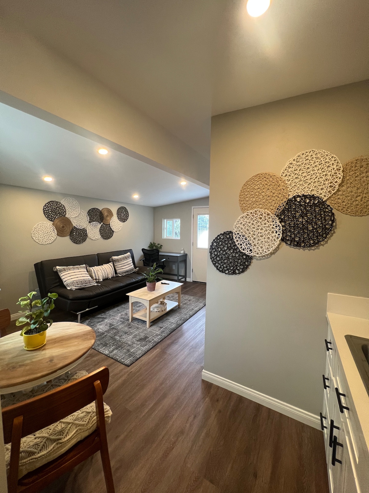 A living area is featured, showcasing a dark sofa with patterned cushions. On the wall, a collection of decorative plates adds visual interest. A small round table is positioned nearby, accompanied by a wooden chair. Natural light is provided through windows, enhancing the inviting space.