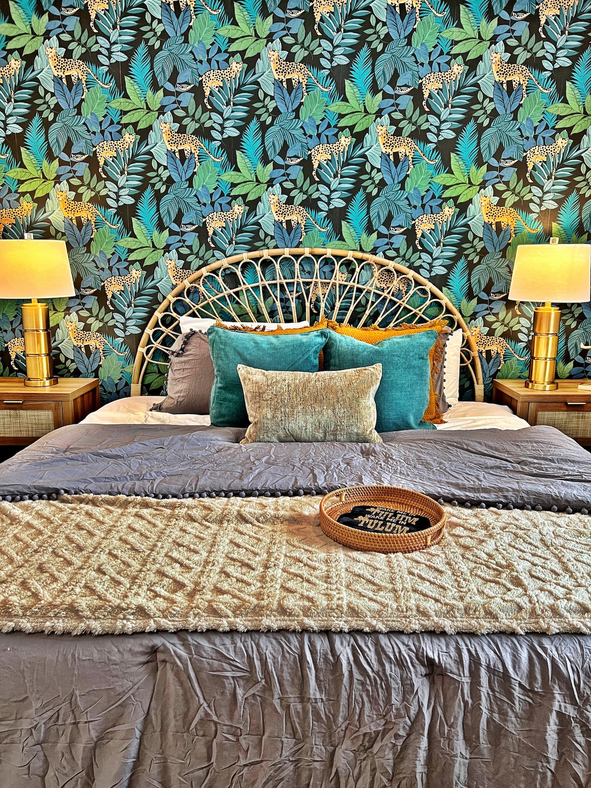 A vibrant bedroom showcases a tropical-inspired wallpaper featuring lush greenery and decorative leopards. The spacious bed is adorned with various pillows in shades of teal and gray. Golden lamps flank the bed, providing warm lighting, while a textured throw blanket adds depth to the inviting design.