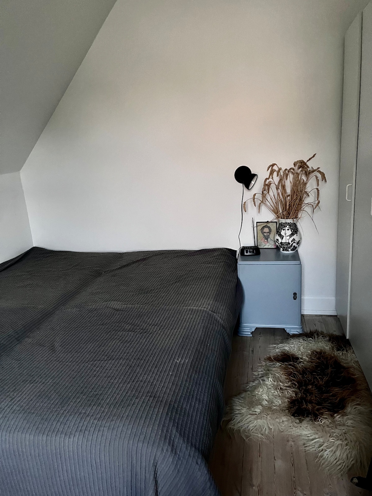 A simple bedroom is presented with a spacious bed covered in a dark striped blanket. A nightstand holds a small vase with dried flowers, alongside a framed picture. Soft textures are provided by a furry rug on the wooden floor, complementing the minimalist decor.