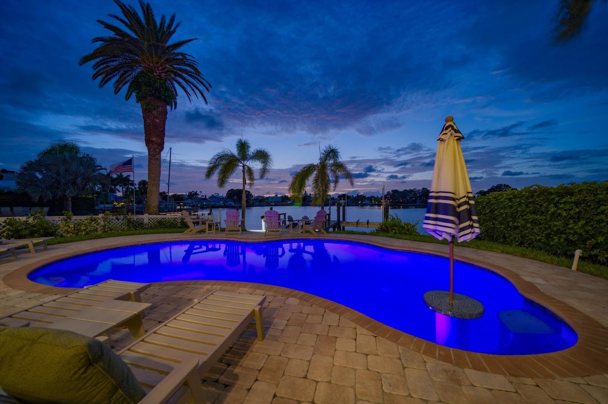 The outdoor area features a heated saltwater pool illuminated with a soothing blue hue. Surrounding palm trees and tropical landscaping enhance the tranquil atmosphere. A yellow and white striped umbrella provides shade beside the pool, offering a picturesque view of the waterway beyond.