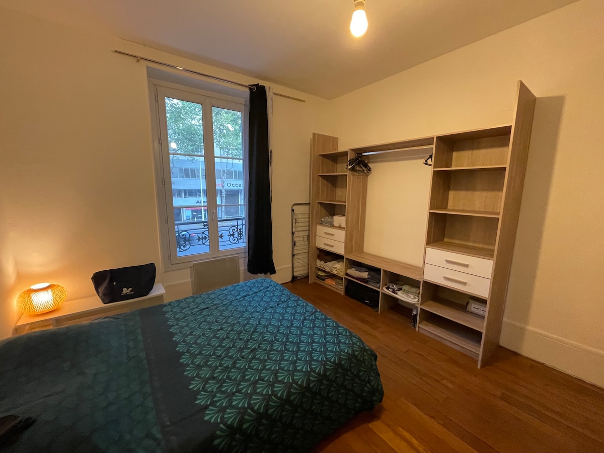 A room features a spacious bed adorned with a green patterned blanket. Natural light enters through a large window dressed with black curtains. A wooden wardrobe includes shelves and hanging space. A small bedside table is positioned beside the bed.