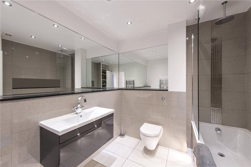 A modern bathroom features a large vanity with a sleek sink and a toilet. A glass shower is visible alongside a bathtub. The space is well-lit with recessed ceiling lights, and mirrored surfaces enhance the sense of openness.