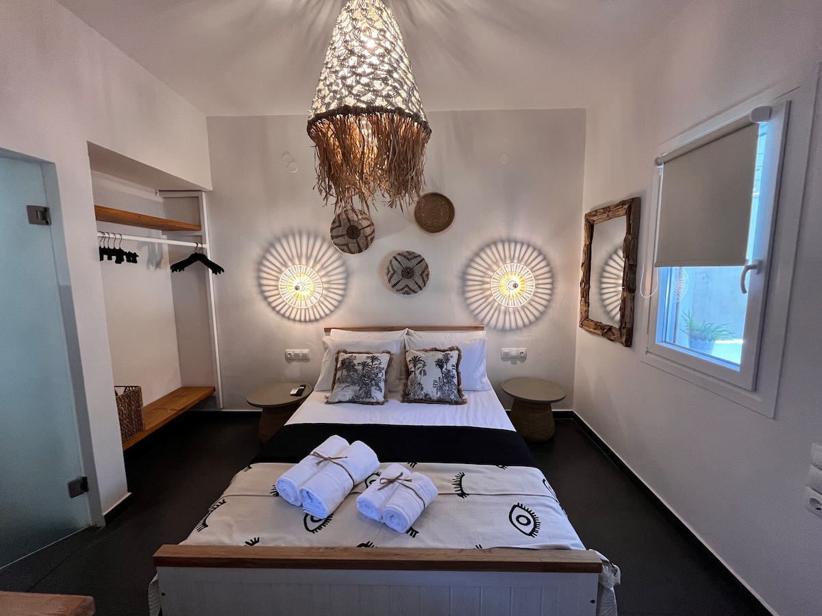 A modern bedroom features a spacious bed adorned with a patterned throw and neatly arranged towels. Light fixtures with a circular design flank the bed, while a minimalist wardrobe and decorative wall accents complement the neutral-colored walls. Natural light enters through a window covered by light shades.