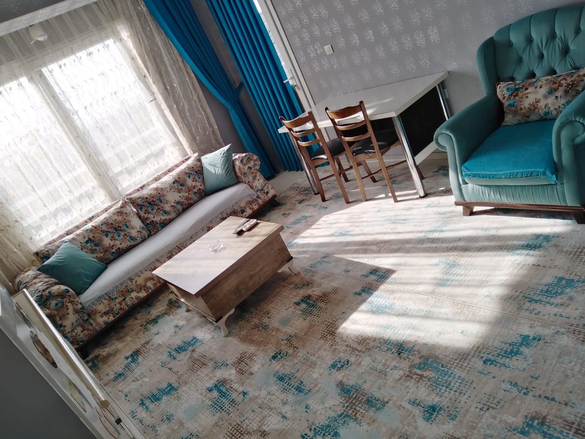 A living area is presented with a floral patterned sofa and a coordinating armchair. A wooden coffee table sits at the center, while a dining table with chairs is positioned nearby. Light filters through sheer curtains, creating a warm ambiance.