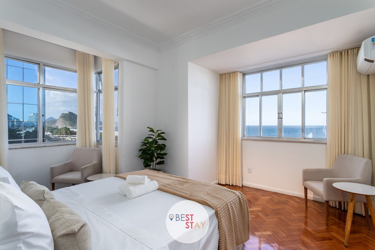 A bright bedroom features large windows that provide stunning views of Copacabana beach. Natural light fills the space, complemented by light-colored curtains. The room includes a spacious bed with white linens, a small round table, and two armchairs, creating a comfortable atmosphere.