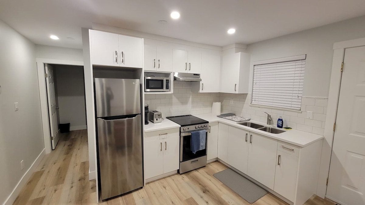 A modern kitchen is featured with stainless steel appliances, including a refrigerator, microwave, and gas stove. White cabinetry contrasts with light wood flooring. Natural light enters through a window, and a small mat lies in front of the sink area.