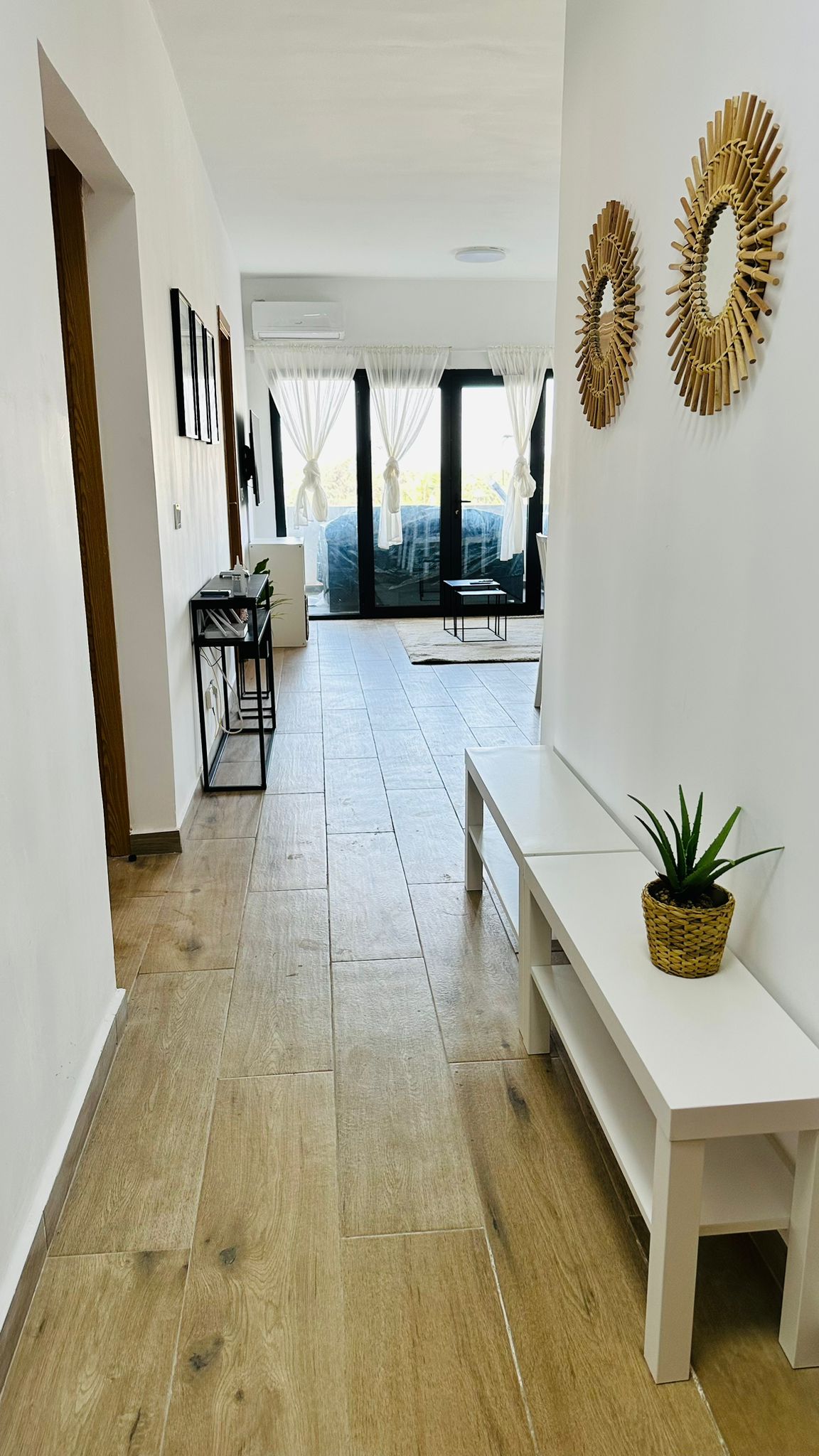 A hallway is presented with light-colored walls and a tiled floor, leading to a bright living area. A narrow white bench and a small potted plant are visible on one side. Sunlight filters through large glass doors at the end of the hallway.