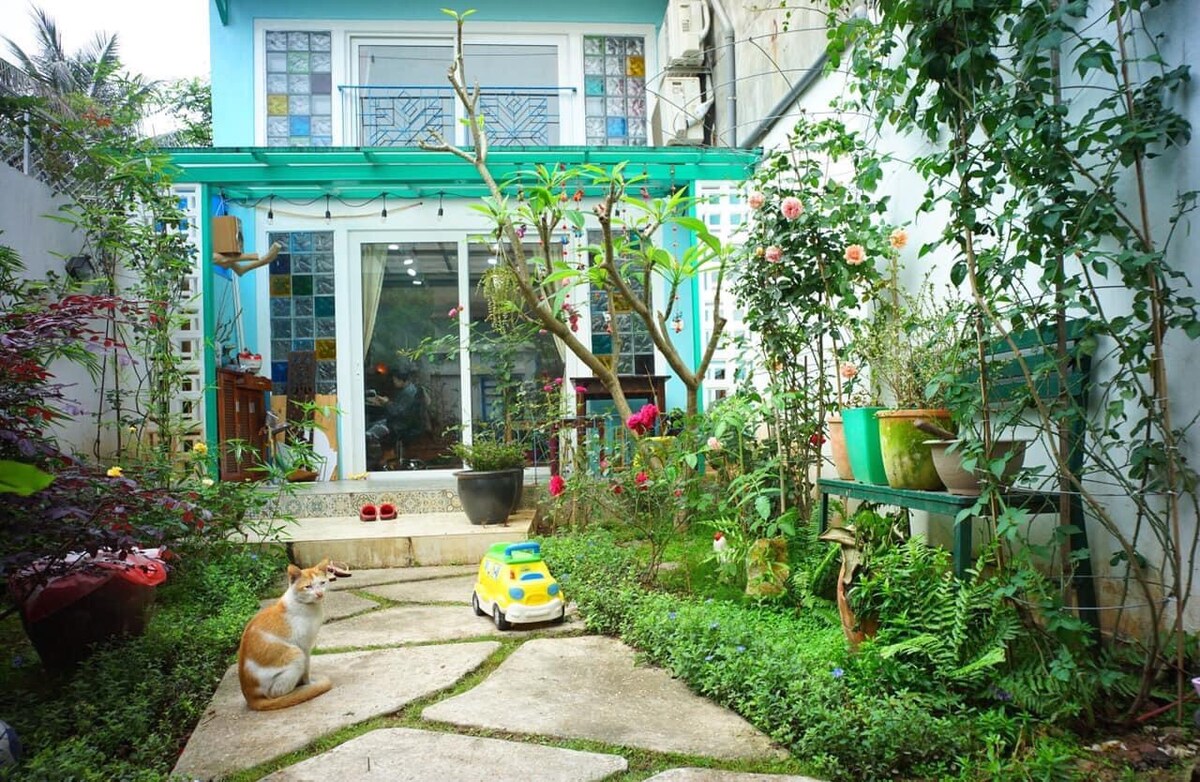 A charming garden space is visible in front of a two-story house, featuring a path lined with greenery and colorful flowers. A small, playful dog sits on the stone pathway, adding a lively touch to the serene environment. Large glass doors offer a glimpse into the indoor area.
