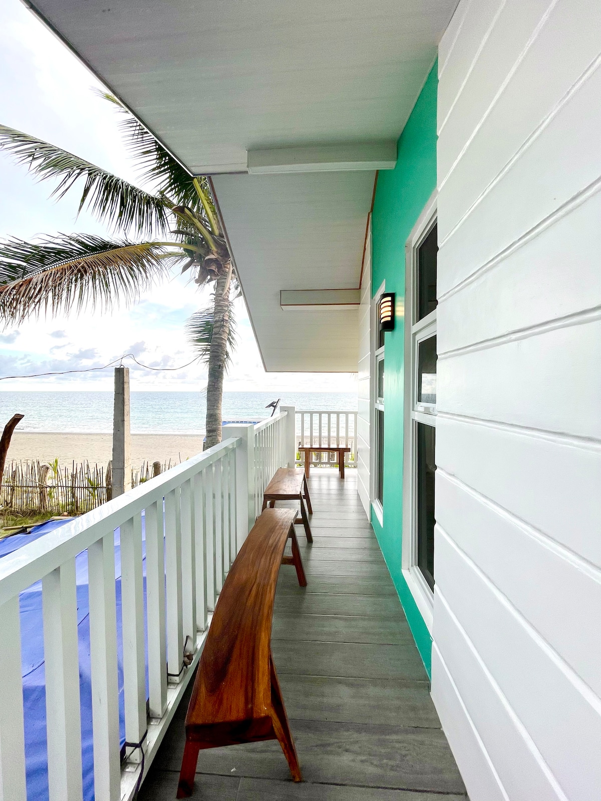An outdoor balcony features wooden benches along a white railing, with views of the beach and palm trees. The turquoise-colored wall provides a vibrant contrast to the white exterior, and natural light filters in from the open space.