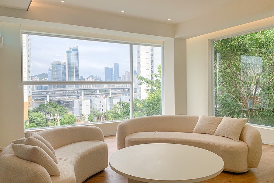 A spacious living area is showcased with large windows offering expansive views of the city skyline. Two curved, soft sofas are positioned around a round coffee table, inviting relaxation and conversation. Natural light fills the room, highlighting the neutral color palette and modern design.