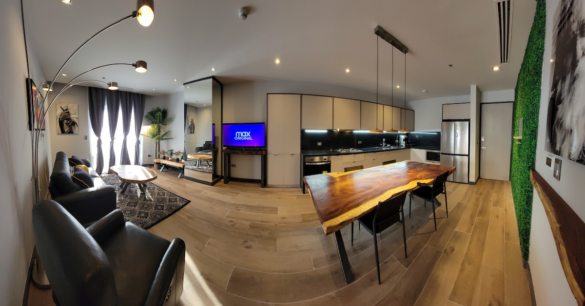 A contemporary living space is presented, featuring a spacious dining area with a unique wooden table. Comfortable seating is arranged in the lounge area, while large glass doors provide a view into a fully equipped kitchen. Natural light enhances the modern design.