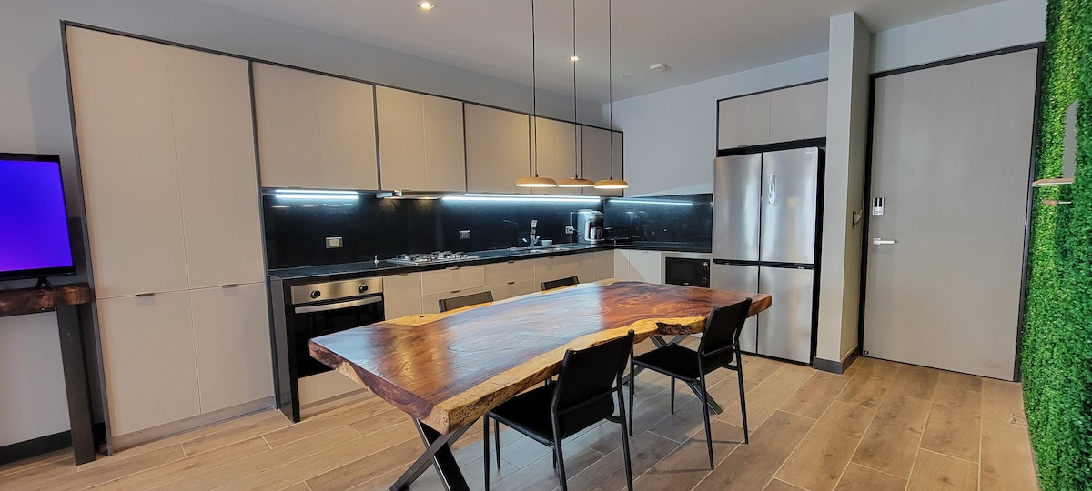 A modern kitchen area features a large wooden dining table surrounded by four black chairs. Sleek cabinetry is complemented by stainless steel appliances, including a refrigerator and stove. Soft lighting accentuates the natural wood grain of the table and the minimalist design.