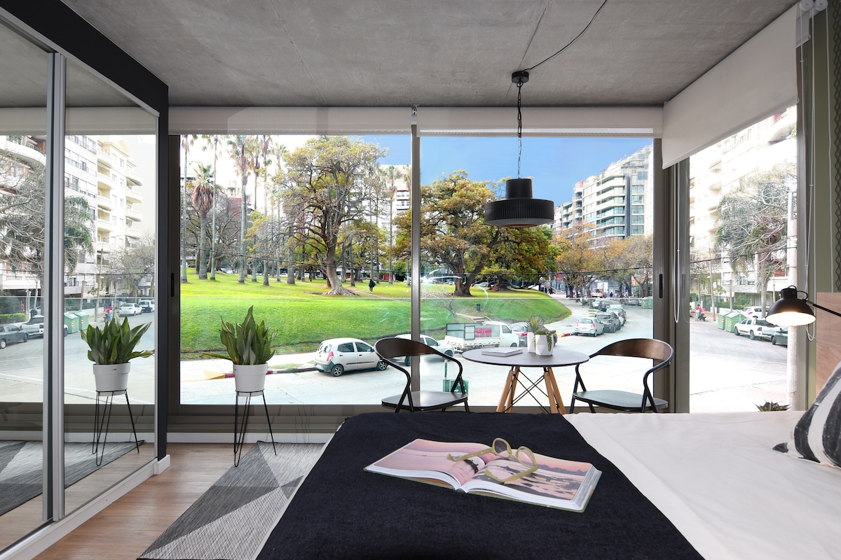 A spacious studio features floor-to-ceiling windows offering views of Biarritz Park. Two chairs and a small table are positioned near the window, inviting relaxation. The room is furnished with a comfortable bed dressed in dark linens, creating a cozy atmosphere with natural light filtering through.