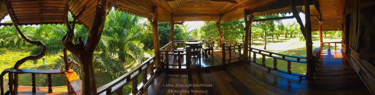 A spacious wooden terrace features a large table surrounded by chairs, overlooking lush palm trees. The wooden flooring highlights a natural aesthetic, providing an inviting space for family gatherings. The open layout enhances air circulation and offers a view of the surrounding garden.