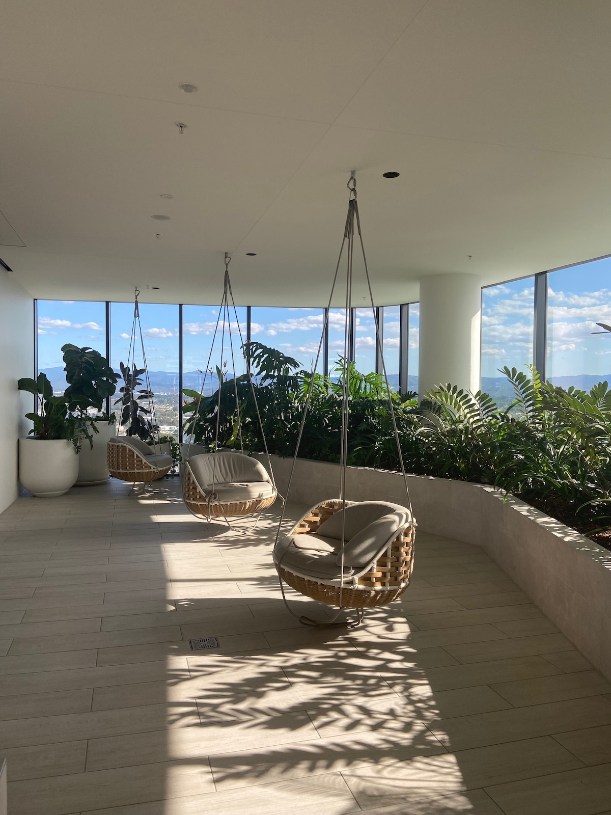 A bright, airy space features three hanging chairs with soft cushions, positioned along a floor-to-ceiling window. Lush greenery adorns the surroundings, creating a serene environment filled with natural light and offering views of the outdoors.