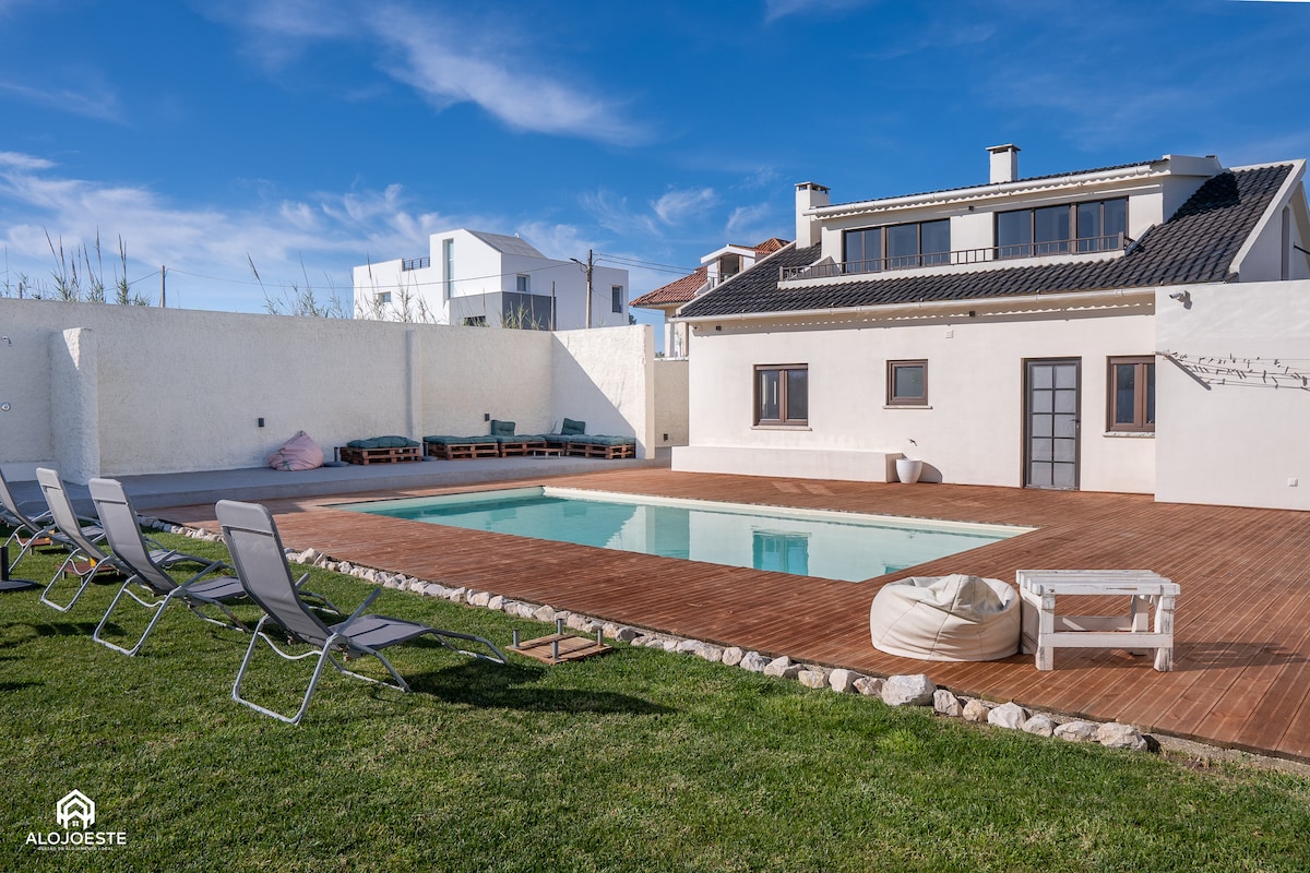 A spacious outdoor area features a well-maintained swimming pool surrounded by wooden decking. Lounge chairs are arranged along the pool's edge, with a grassy area nearby. The two-story house is visible in the background, equipped with multiple windows that allow natural light to enter.