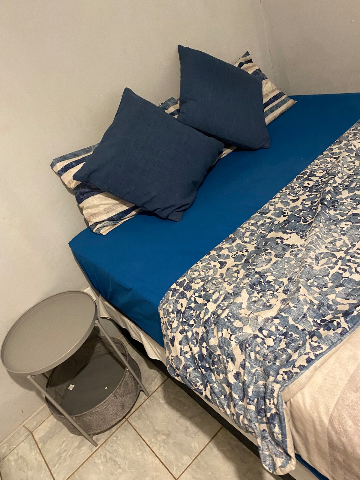 A neatly arranged bed features a blue bedspread complemented by patterned pillows in varying shades. A small side table, crafted in a minimalist design, rests beside the bed, enhancing the room's functionality.
