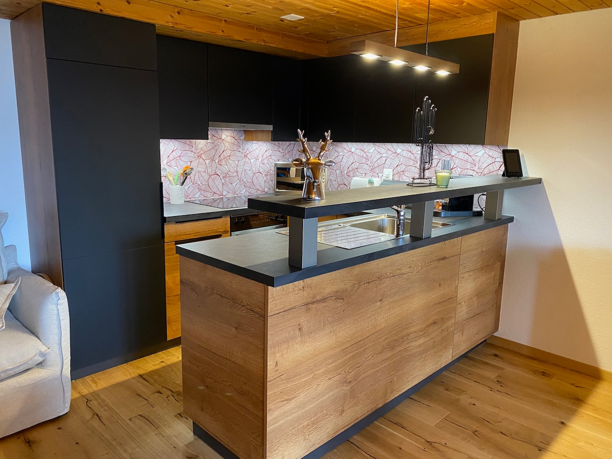 A modern kitchen features dark cabinetry combined with wooden accents, creating a sleek appearance. A countertop with seating is present, alongside essential appliances such as a stove and oven. Decorative elements, including a patterned backsplash, contribute to the inviting design.