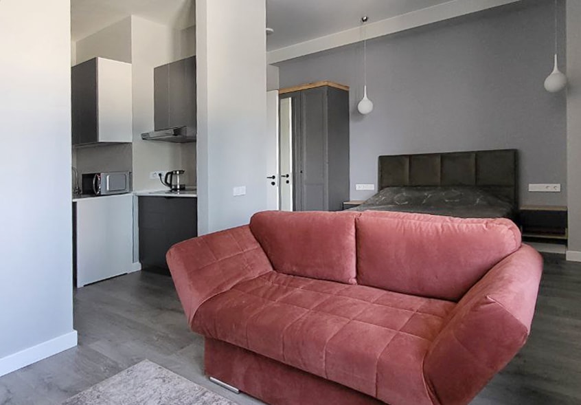 A modern studio apartment is presented with a cozy sofa positioned in the foreground. The kitchenette is located in the background, and a spacious bed is visible to the side, providing a sense of an open layout. Light-colored walls enhance the feeling of space.