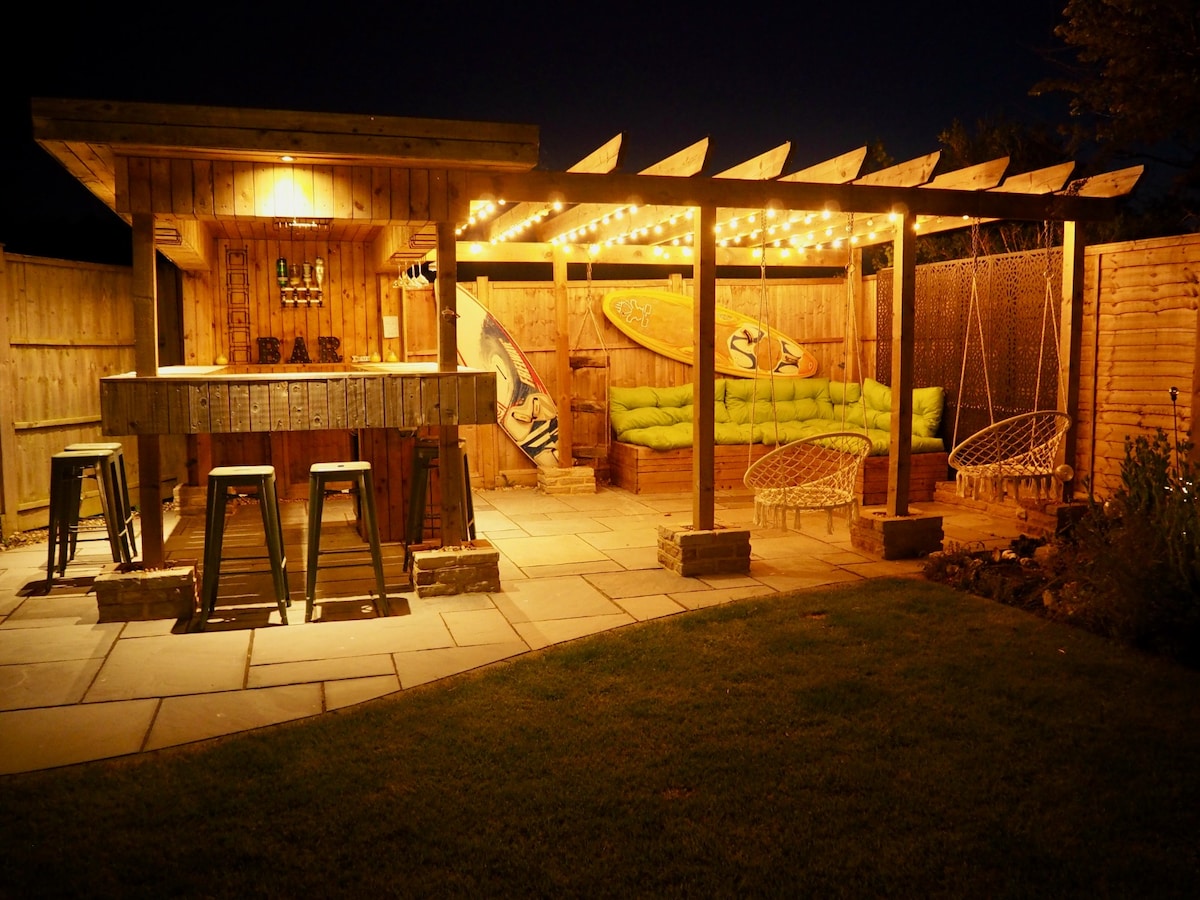 An outdoor bar area is illuminated by string lights, featuring a wooden bar with seating for four. Comfortable seating is arranged around the space, accented by surfboard decorations. A stone patio provides a spacious area, complemented by surrounding mature landscaping.