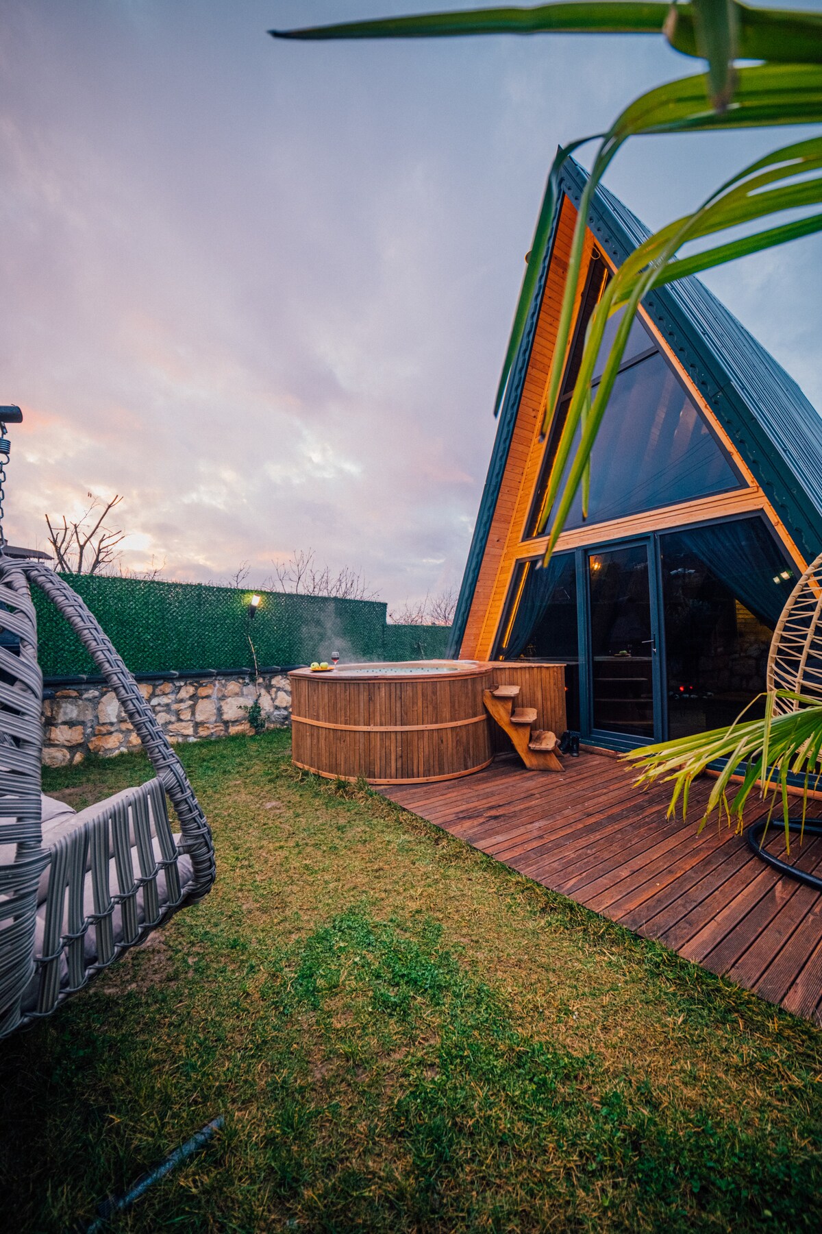 A modern A-frame bungalow is surrounded by a well-maintained garden, featuring a wooden mini pool on a deck. A swing is positioned nearby, and the structure's large windows invite natural light. Soft hues of sunset create a serene ambiance in the background.
