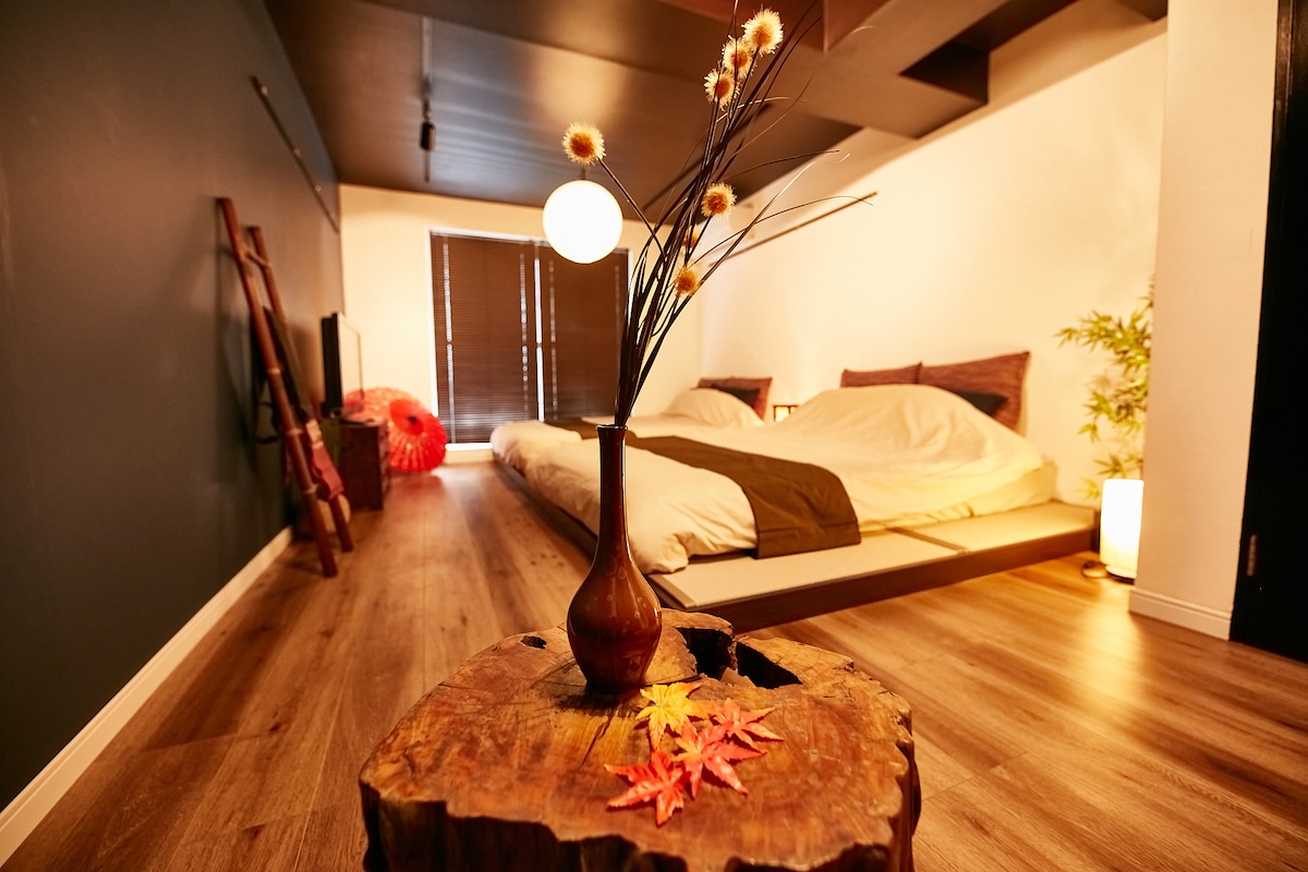 A spacious room features a traditional Japanese bed set up with neutral bedding. A wooden table displays decorative flowers and vibrant autumn leaves. Soft lighting from lamps creates a calm environment, complemented by dark walls and natural wooden flooring.