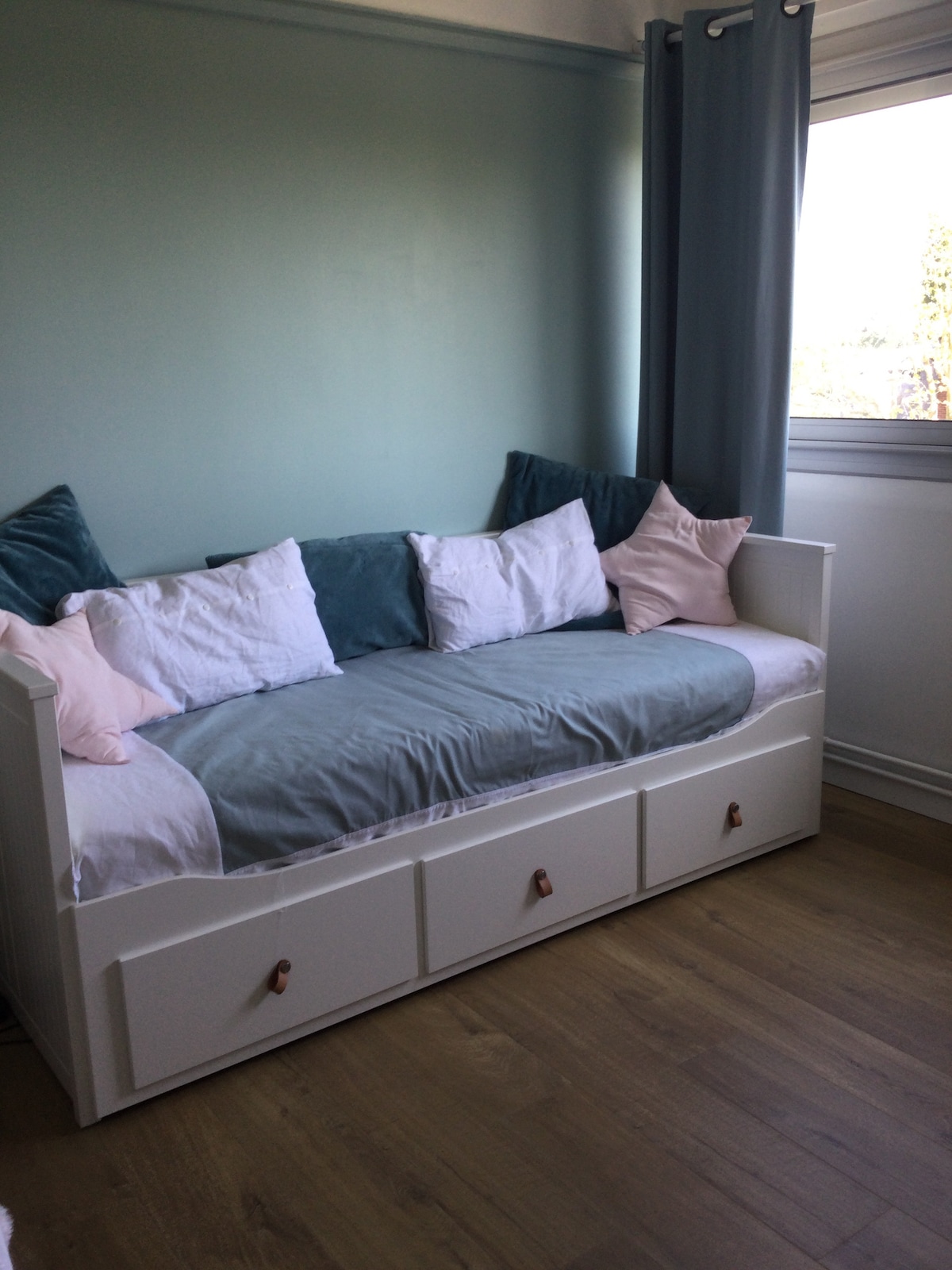 A well-appointed corner features a daybed with a light-colored frame. Decorative pillows in soft tones are arranged along the back, enhancing comfort. Natural light enters through the window, illuminating the adjacent greenery outside.