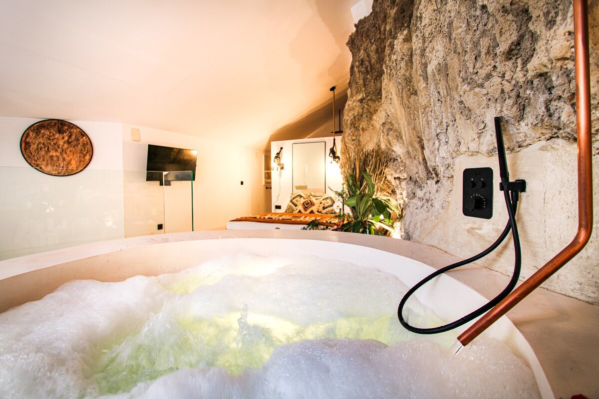A private, heated jacuzzi is featured in a serene setting, surrounded by natural stone walls. Soft bubbles gently rise to the surface, while warm lighting creates a soothing ambiance. A glimpse of the bedroom area is visible in the background, enhancing the tranquil atmosphere.