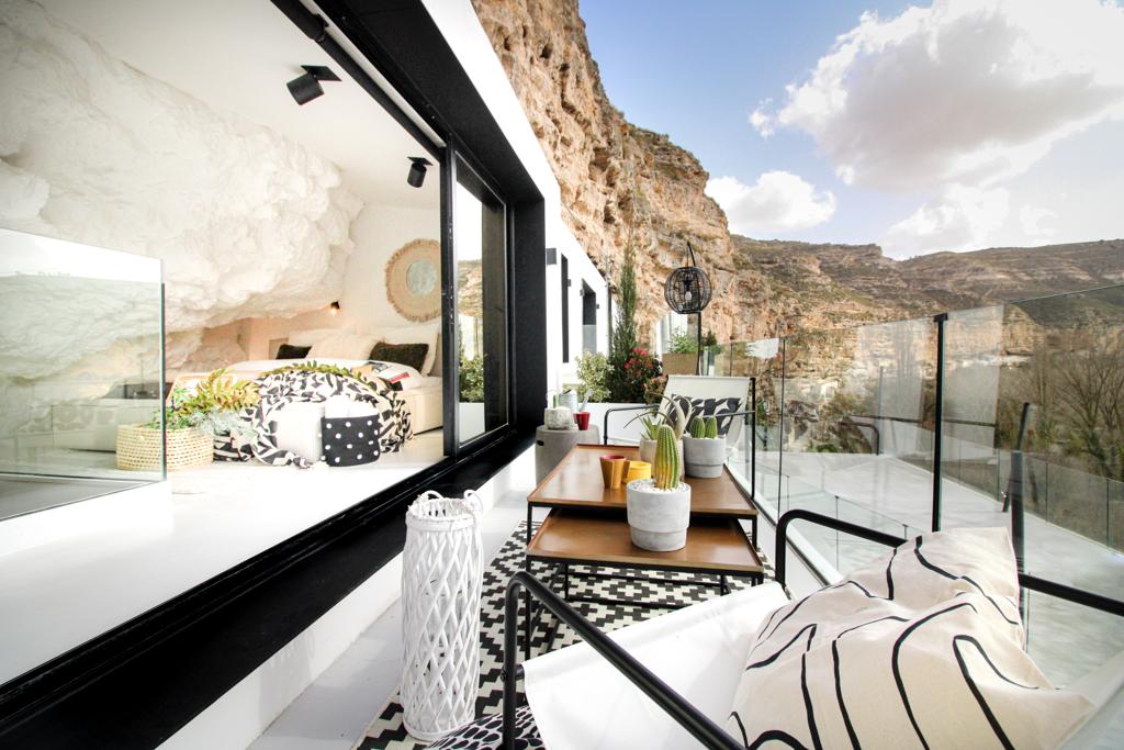A terrace offers a scenic view of the surrounding rocky landscape, furnished with a small table and comfortable seating. Large glass doors lead into a cozy bedroom area, featuring natural stone elements and minimalist decor. The contrast of materials creates a harmonious blend with nature.