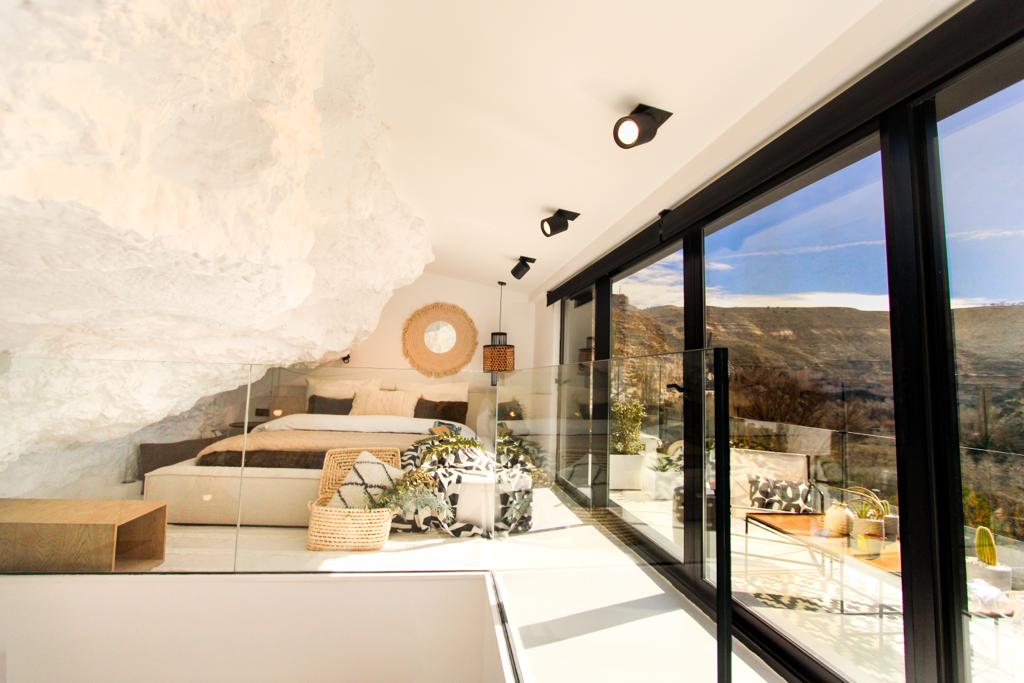 A spacious bedroom features a large King-size bed set against natural rock walls. Floor-to-ceiling glass doors provide panoramic views of the valley, complemented by modern lighting fixtures. A serene atmosphere is created with minimalist decor, including a textured rug and indoor plants.