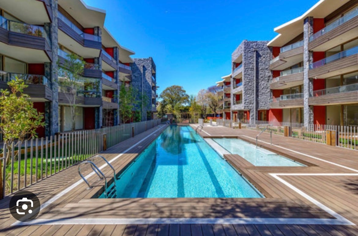 A central swimming pool is flanked by modern apartment buildings, featuring a wooden deck and landscaped greenery. The pool is surrounded by a secure fence, with lounge areas and sun loungers visible along the edges.