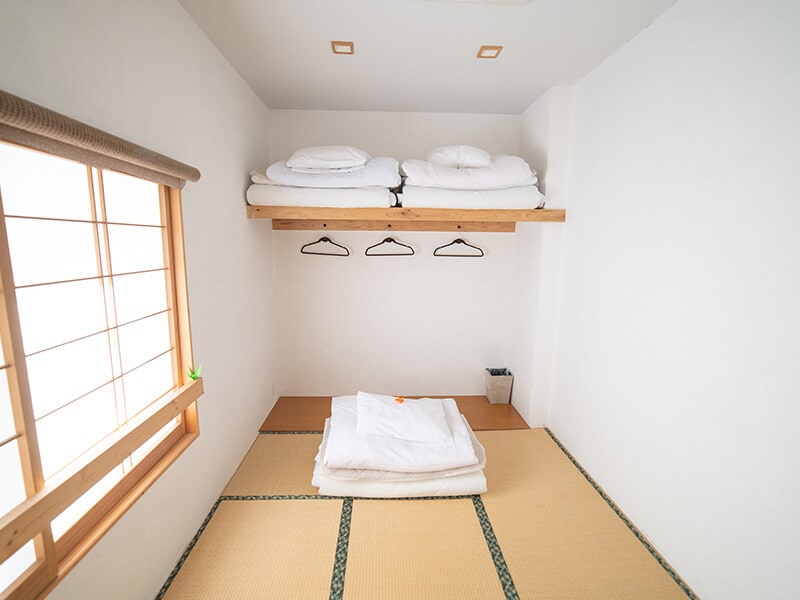 A minimalist room features traditional tatami flooring, with a spacious sleeping area. A lofted bed with neatly arranged bedding sits accessible by wooden shelving. Natural light filters through a window adorned with wooden slats, enhancing the room's serene atmosphere.