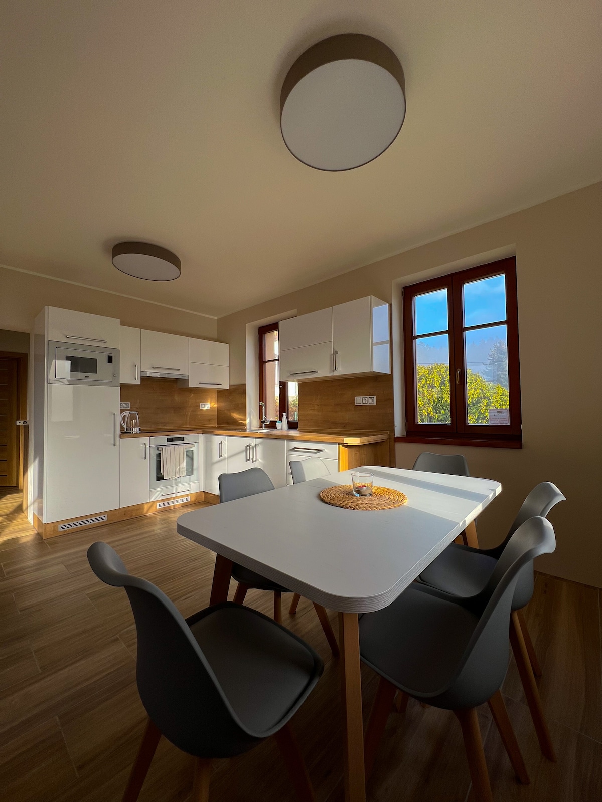 A modern kitchen is showcased with sleek white cabinetry and wood accents. A round dining table with a woven centerpiece is surrounded by four chairs, with sunlight streaming through two windows that offer a view of the outdoor greenery.
