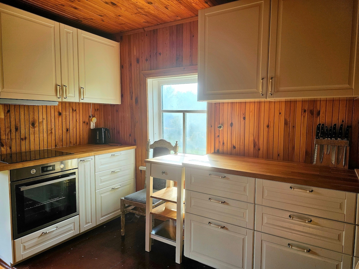 A well-appointed kitchen features wooden cabinetry and a spacious countertop. An oven and a window allow for ample natural light. A chair sits next to a small dining table, complemented by a set of knives displayed on a magnetic strip.