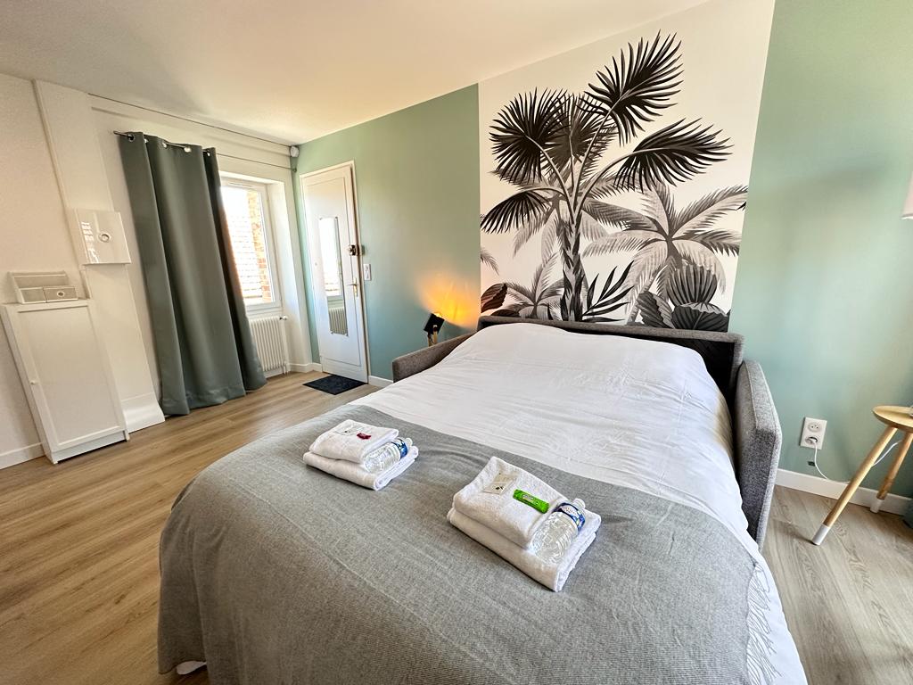 A spacious sleeping area features a large bed dressed in crisp linens, with white towels neatly arranged on top. A wall adorned with tropical plants creates a calming ambiance, while natural light enters through a nearby window, complemented by soft green walls.