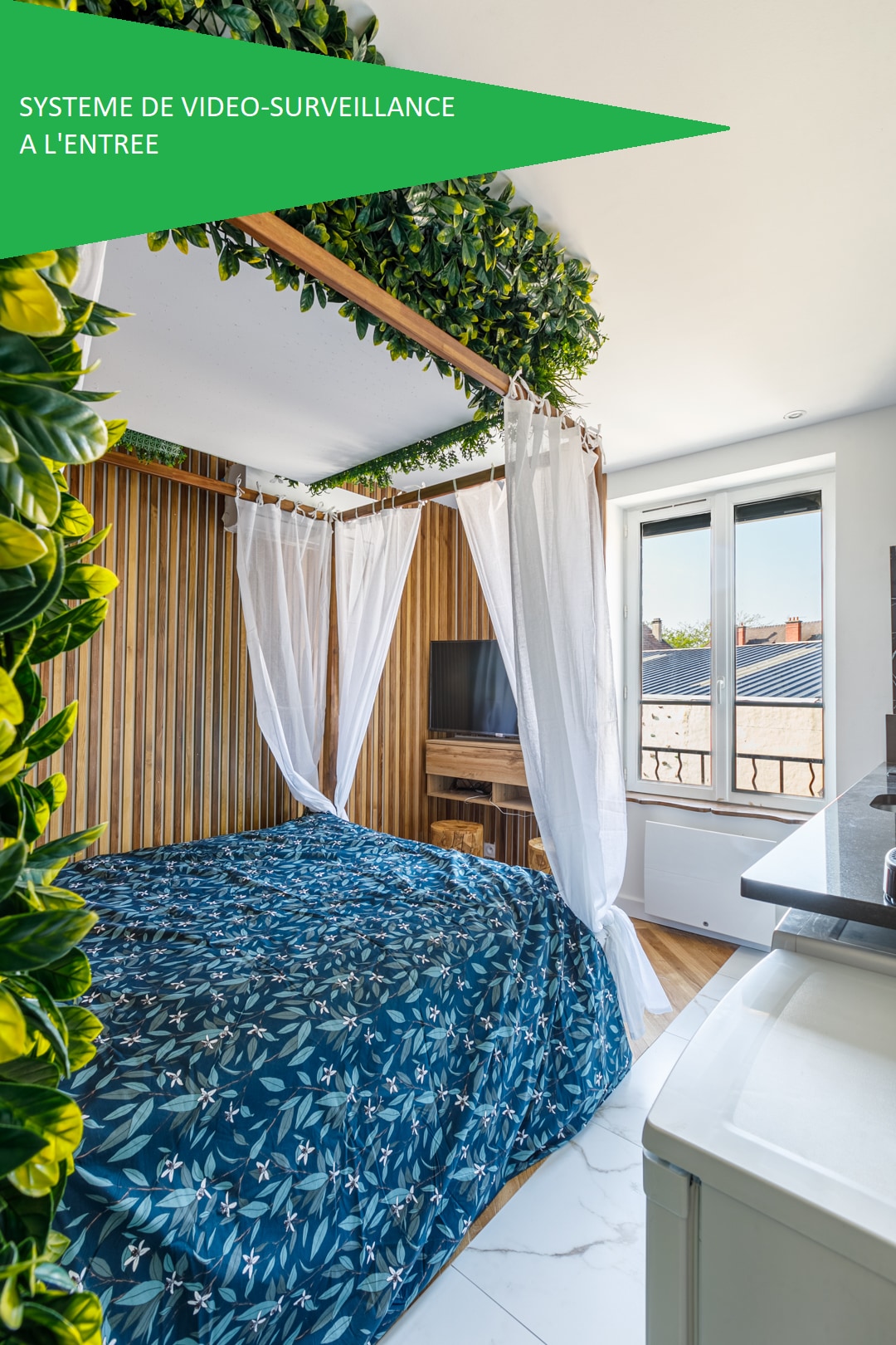 A cozy sleeping area is showcased, featuring a spacious bed adorned with a blue patterned cover. Light drapes and greenery surround the bed, creating a serene space. A window allows natural light to fill the room, while a television is mounted on the wooden paneling.