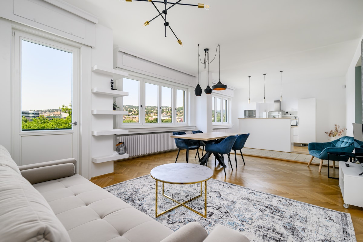 A bright living space is shown, featuring a comfortable sofa and a circular coffee table on a patterned rug. Large windows frame a view of greenery, while a dining area with a table and chairs is visible in the background, along with modern pendant lighting.