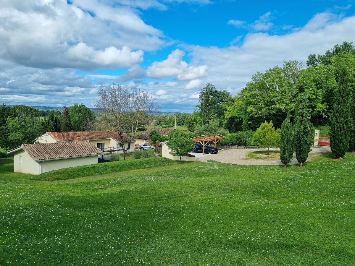 A serene landscape showcases a spacious green lawn framed by trees. Multiple buildings are visible, including a cozy residence and a carport, set against a backdrop of picturesque rolling hills under a partly cloudy sky.