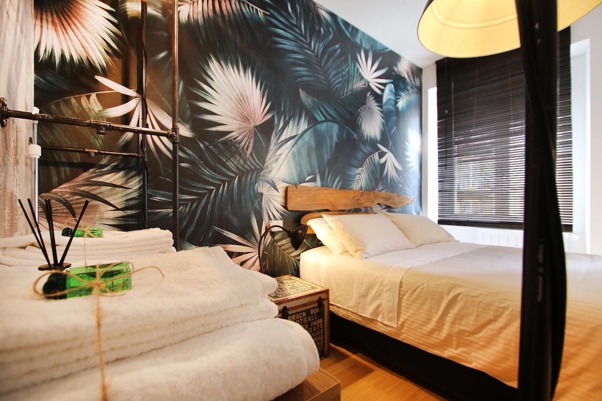 The bedroom features a large bed against a vibrant mural of tropical foliage. Soft, neatly stacked towels are arranged on a nearby unit. Natural light filters through slatted blinds, enhancing the peaceful atmosphere.