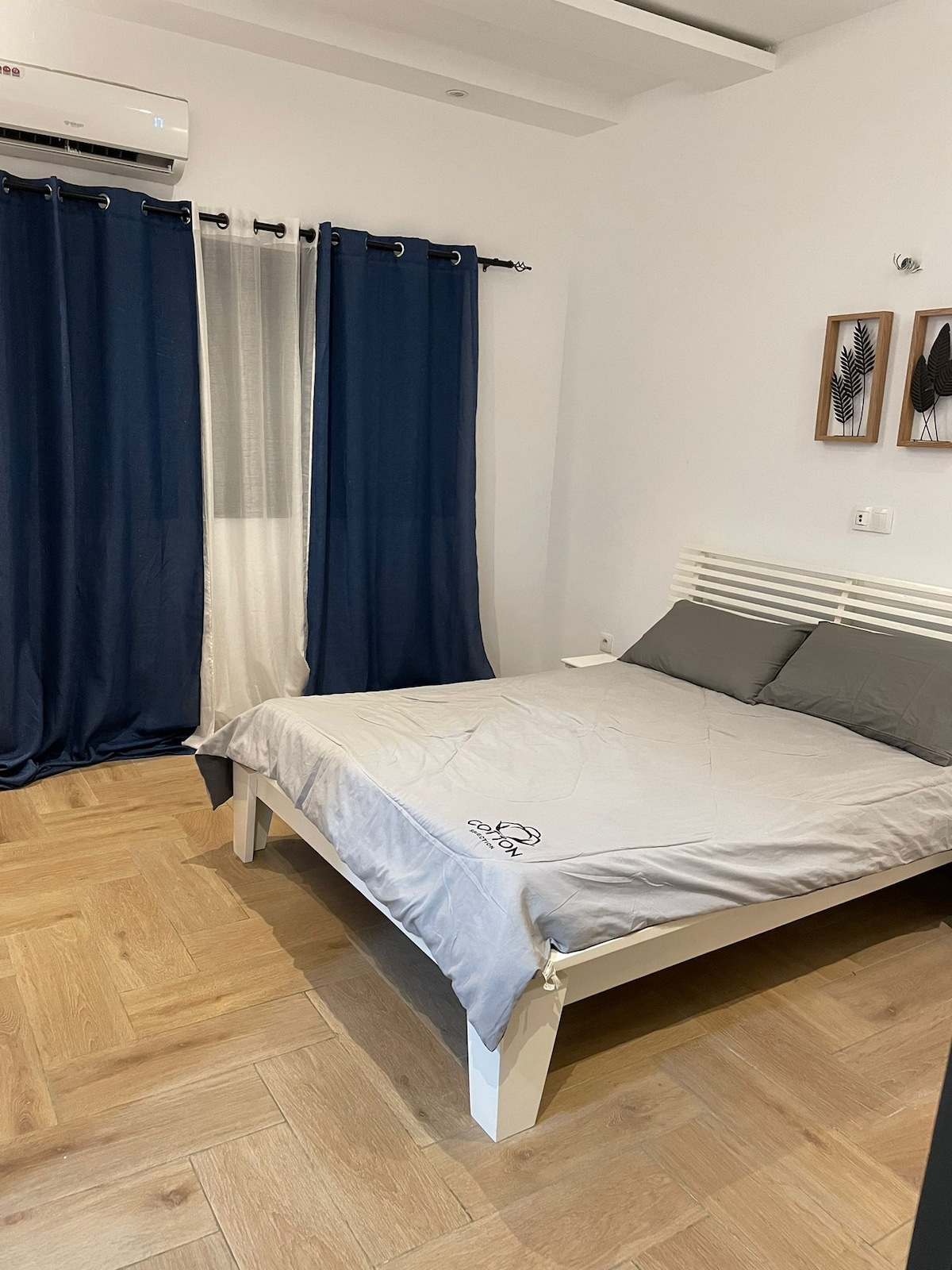 A well-lit bedroom features a spacious bed with gray bedding, complemented by two pillows. Dark blue curtains frame a window, and a wall-mounted light is beside the bed. Natural wood flooring adds warmth to the serene atmosphere.