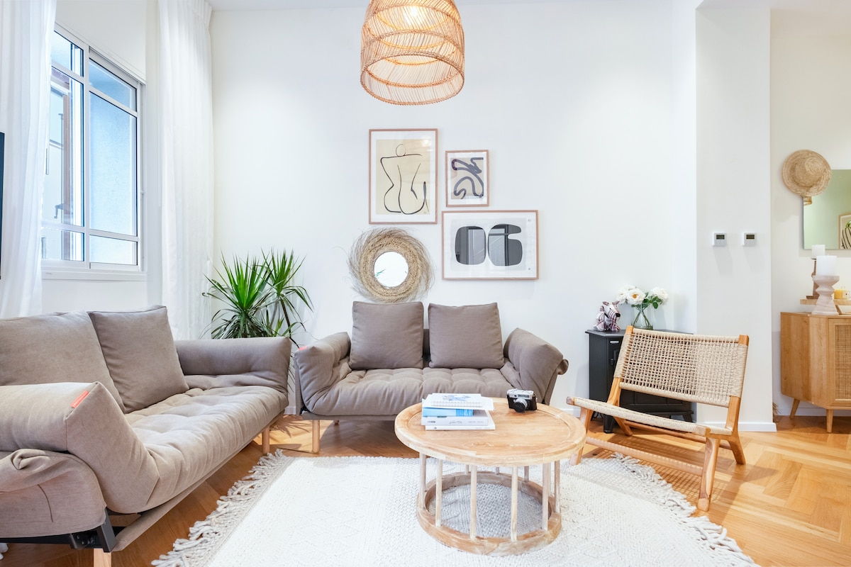 A comfortable living room is highlighted by a large, cozy sofa and a light woven chair. A wooden coffee table sits at the center, surrounded by a soft area rug. Art pieces adorn the walls, and natural light filters through the window, enhancing the welcoming atmosphere.
