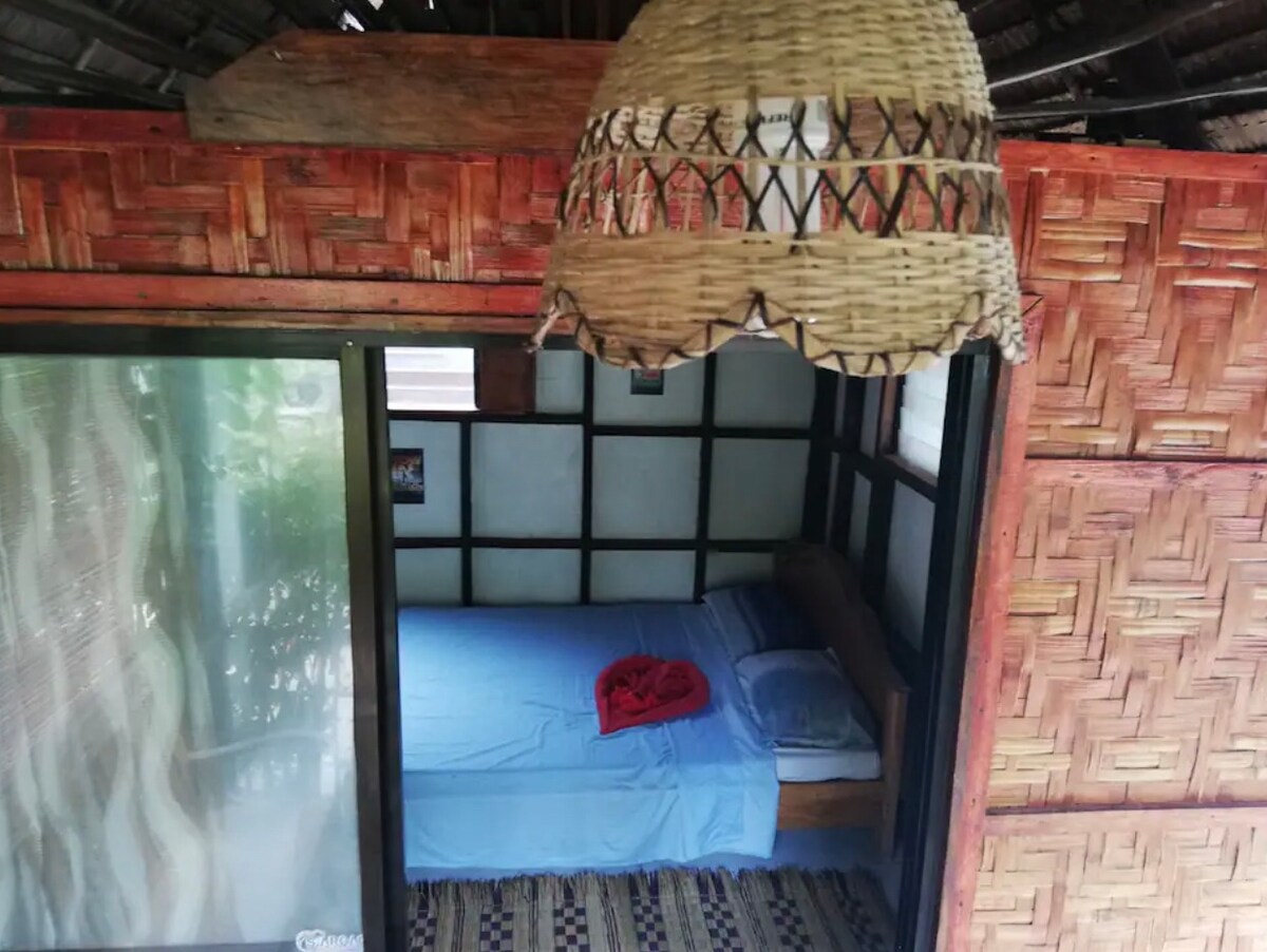 A rustic sleeping area features a wooden frame bed adorned with a light blue blanket and a red throw. A woven ceiling lamp hangs above. The walls are crafted from natural materials, creating a cozy, inviting atmosphere, with a sliding glass door allowing natural light.