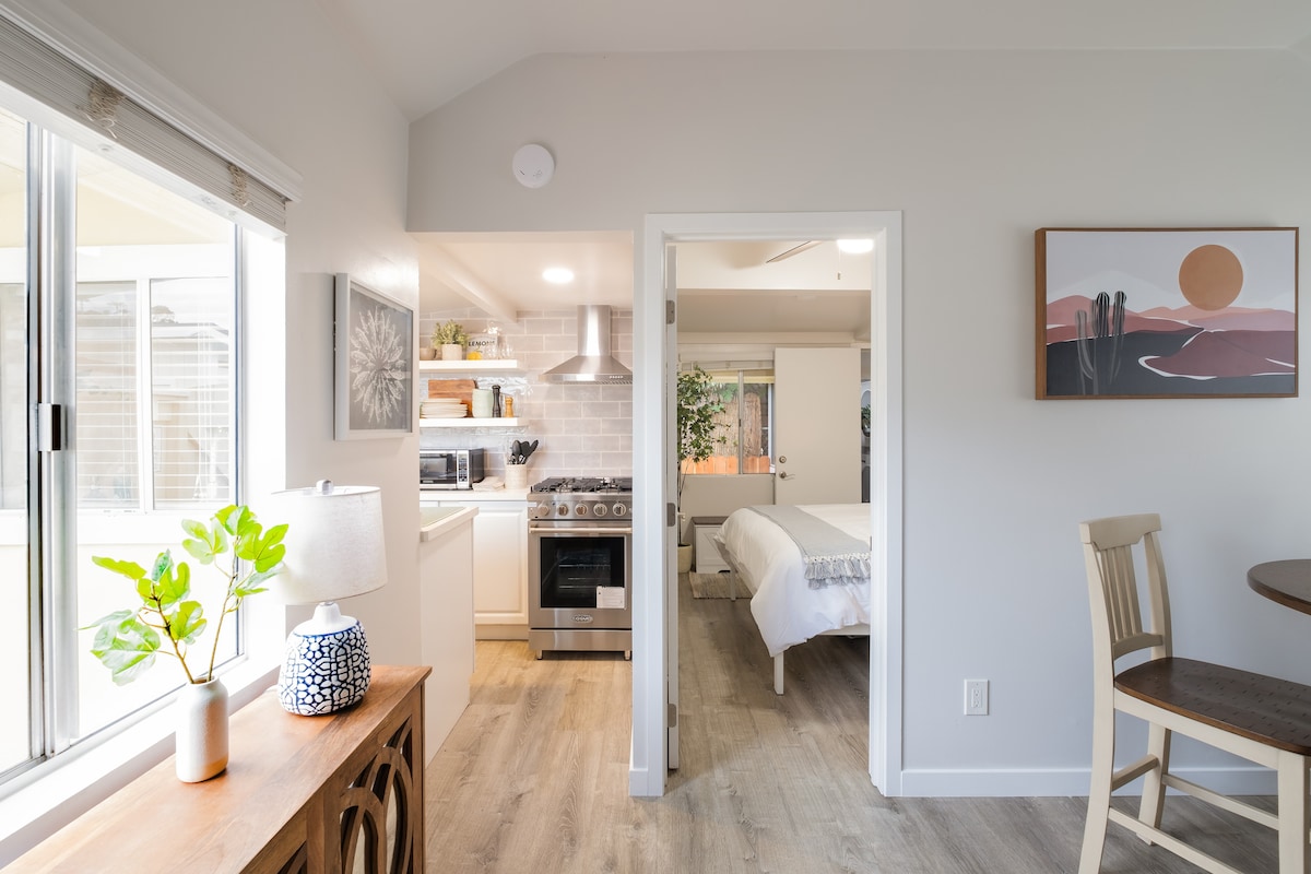 A bright and airy space showcases a modern kitchen area equipped with stainless steel appliances, with natural light streaming in through a large window. A cozy dining table and chair provide a welcoming spot for meals, while a tasteful artwork adorns the wall, complementing the overall design.